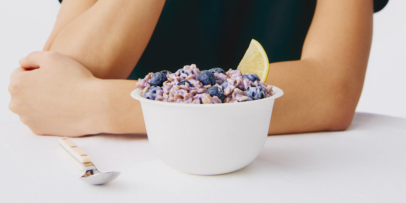 Bowl of cereal with blueberries and a lemon slice on a white surface, person's arms visible.