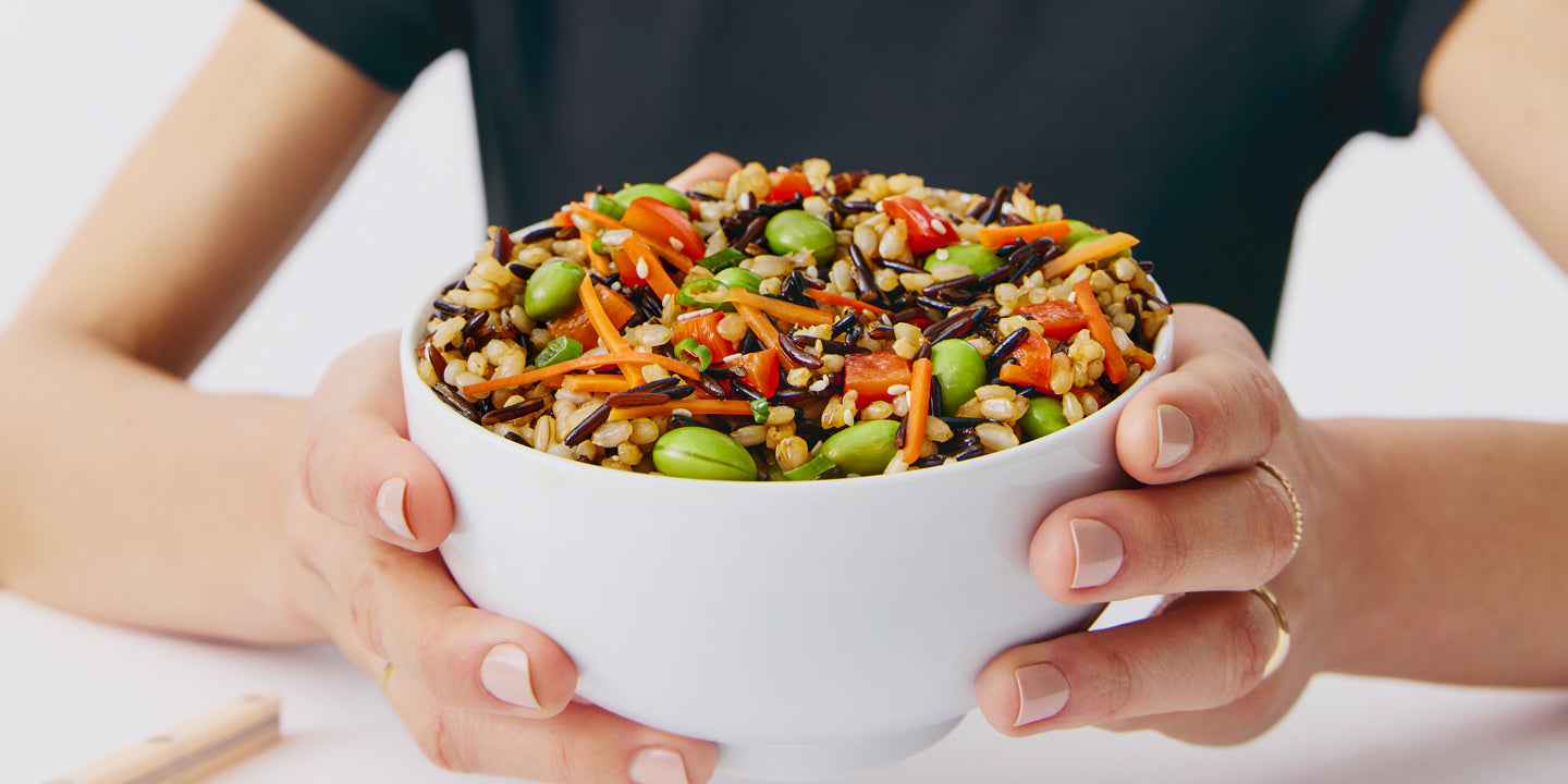 Person holding a white bowl filled with mixed grain salad including edamame, carrots, and wild rice