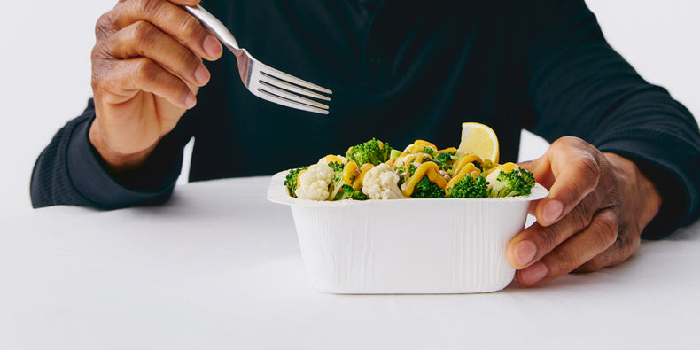 Person holding a white container with broccoli and cauliflower, about to eat with a fork at a white table