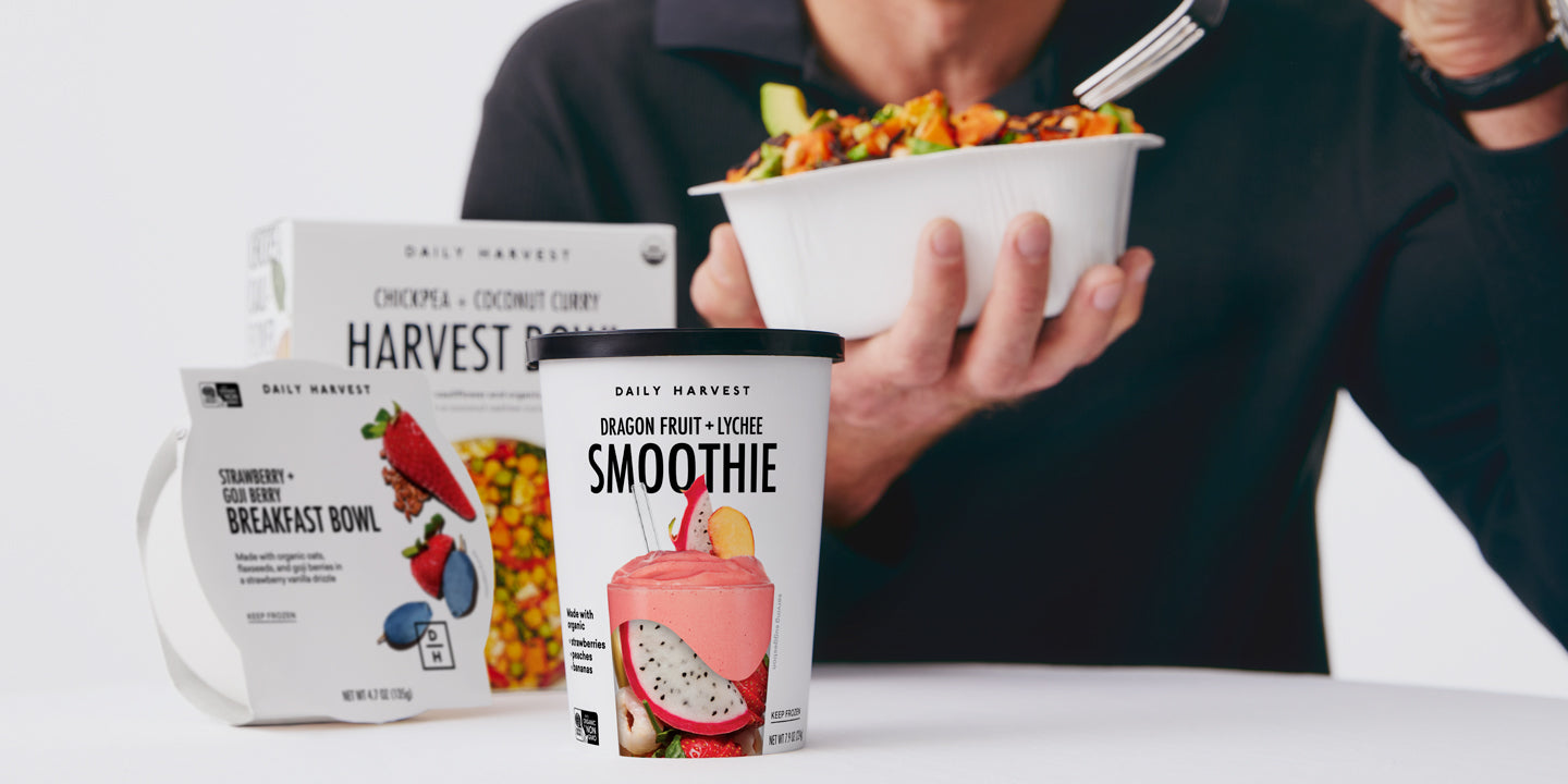 Person eating from a bowl behind various Daily Harvest packaged food items including a smoothie and breakfast bowl