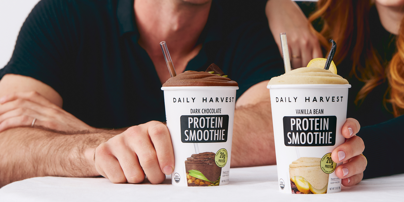 Two people holding Daily Harvest protein smoothies, one dark chocolate and one vanilla bean flavor, on a white surface.