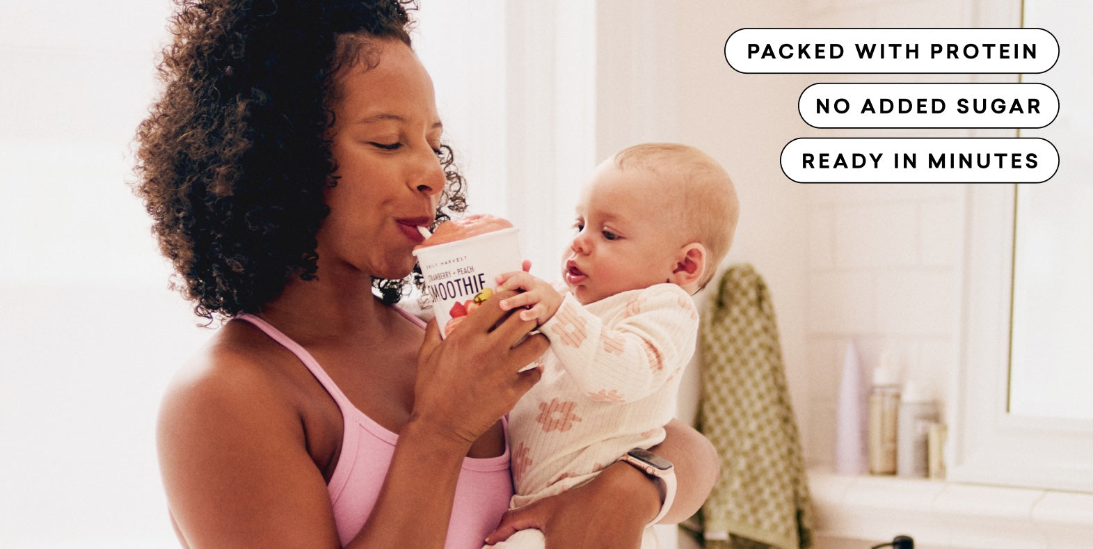 Woman holding a baby and a container of Daily Harvest Smoothie in a bathroom setting.