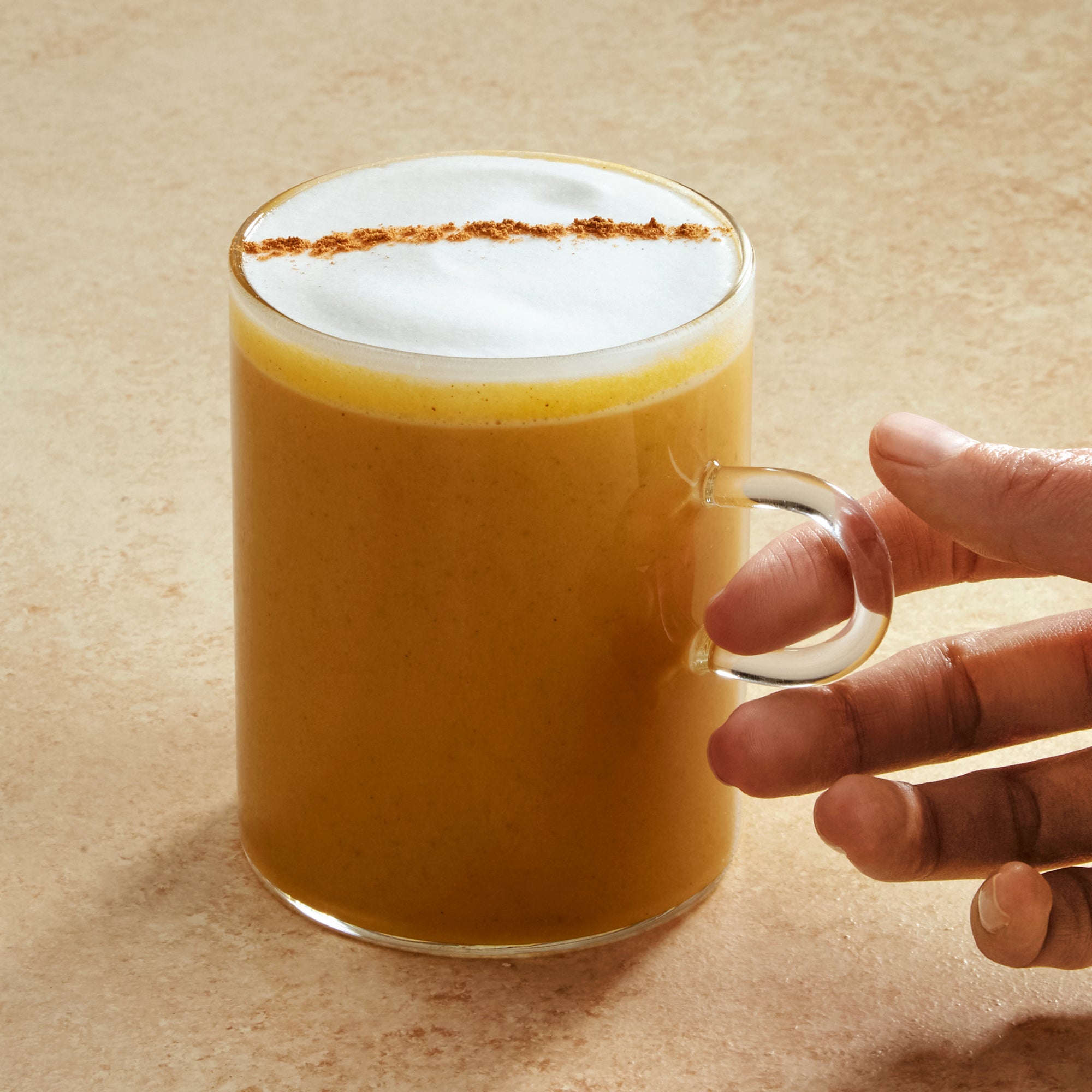 Hand holding a glass of pumpkin spice latte beverage with a frothy top on a beige background