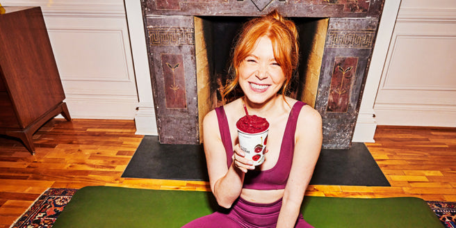 A woman sitting on a yoga mat holding an Acai + Cherry Smoothie with a straw.