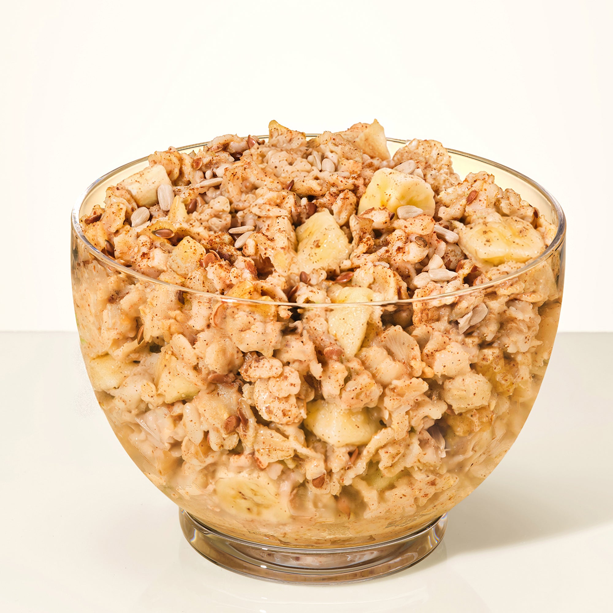 A clear glass bowl filled with a creamy granola or cereal-style snack mix, featuring light brown flakes, pale banana pieces, and small green bits, photographed on a clean white background.