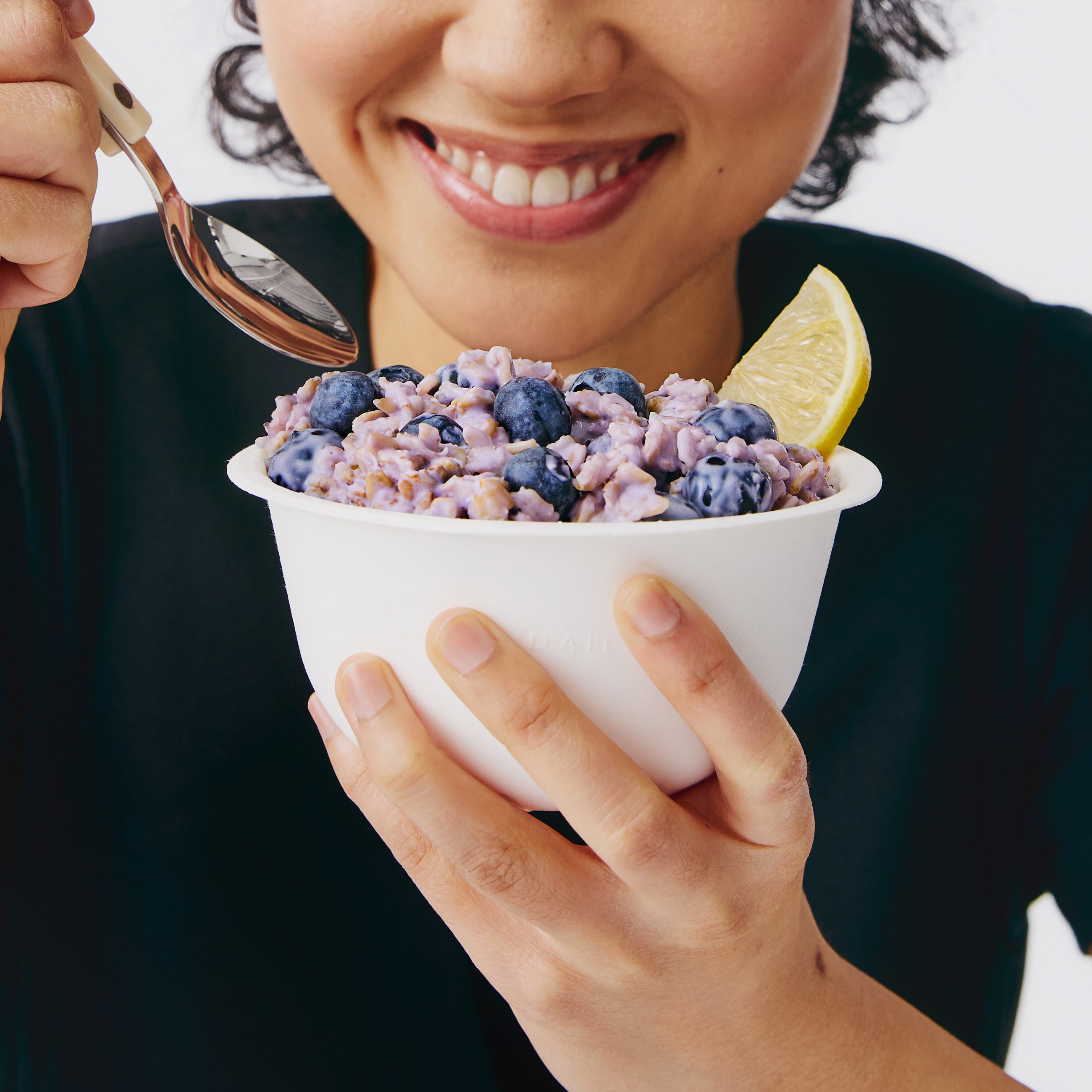 Person holding a bowl of blueberry yogurt with a lemon wedge, smiling.