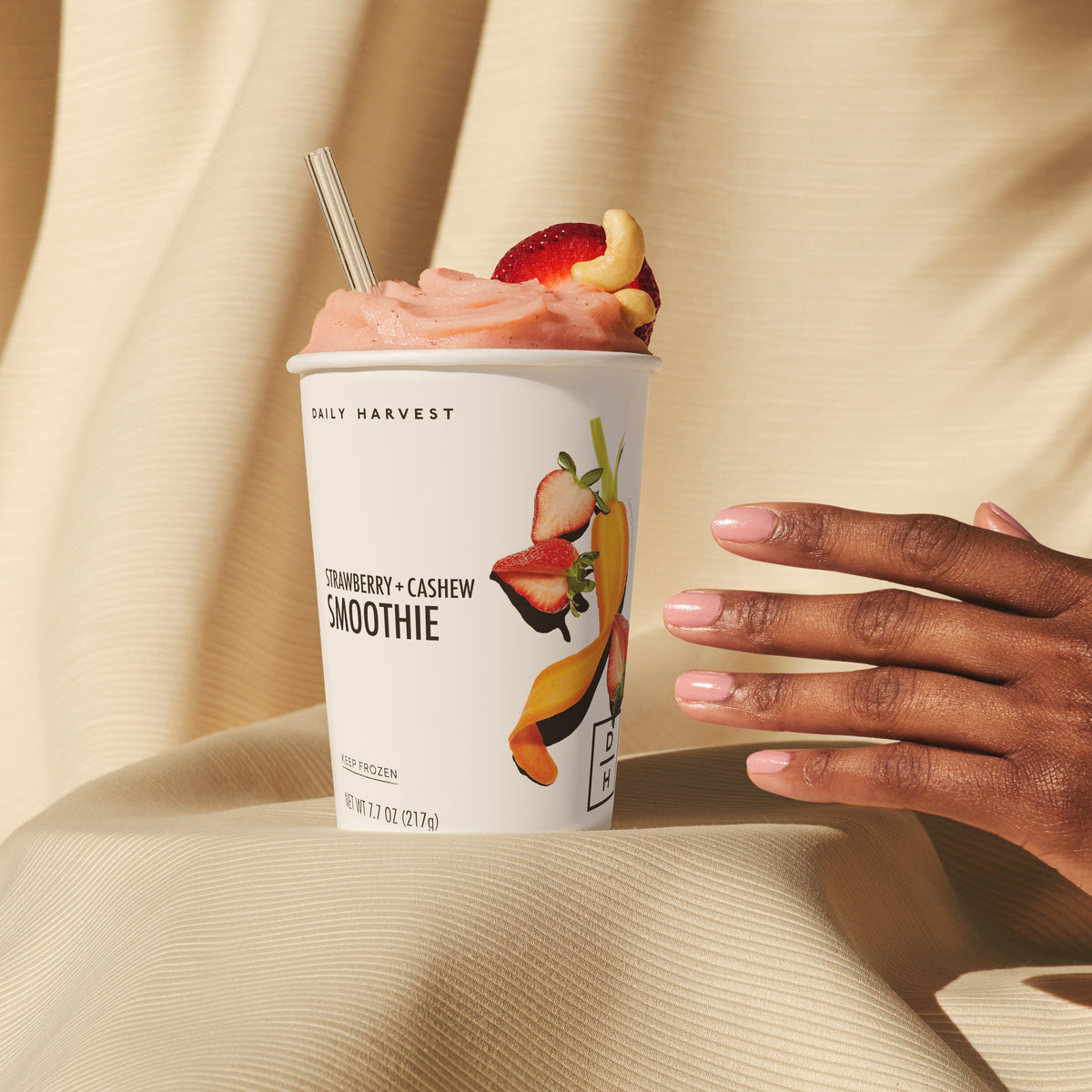 A hand with pink polished nails holds a cup of Daily Harvest strawberry + cashew smoothie with a metal straw, against a beige textured background. The smoothie is pink with fruit pieces on top.